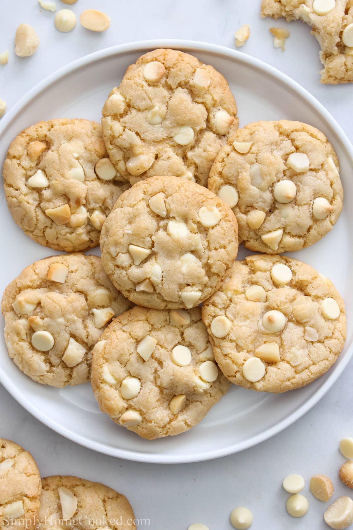 Vertical image of a plate of White Chocolate Macadamia Nut Cookies with nuts and chocolate chips nearby