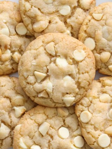 Vertical image of a plate of White Chocolate Macadamia Nut Cookies with nuts and chocolate chips nearby