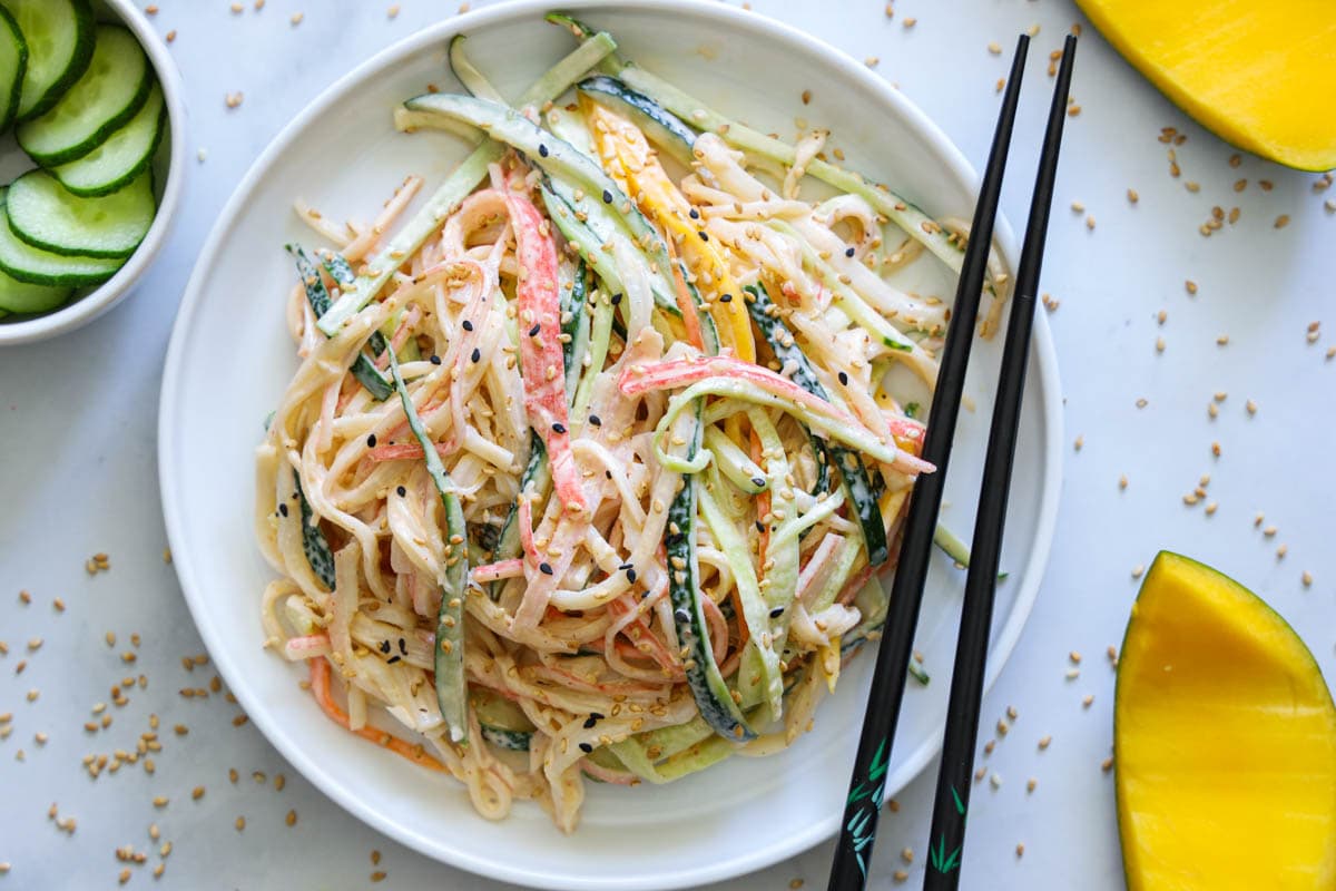 Kani Salad on a white plate with chopsticks and mango and cucumbers on the side