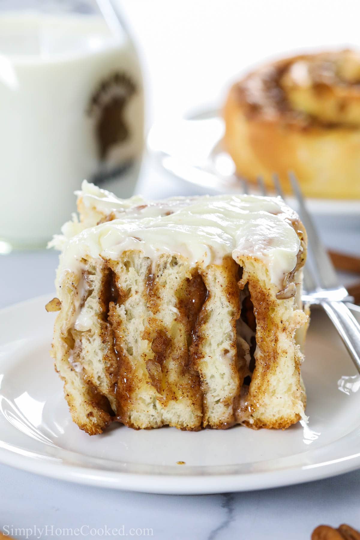 Vertical image of Best Homemade Cinnamon Roll on a white plate with a fork, covered in cream cheese icing