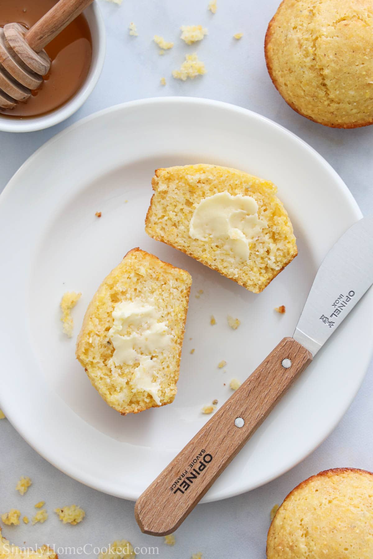 Vertical image of a Cornbread Muffin cut open and buttered on a white plate with a knife and with honey nearby