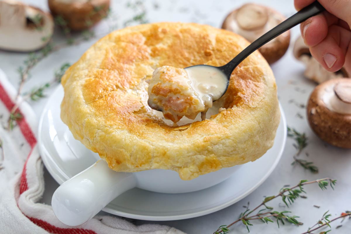 Horizontal image of Cream of Mushroom Soup in a white bowl with a puff pastry top and a spoonful being lifted out