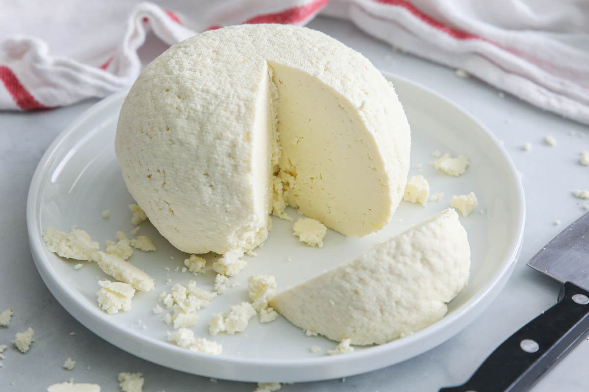 Horizontal image of Farmer's Cheese molded into a ball and a slice cut out, a knife and cheesecloth nearby