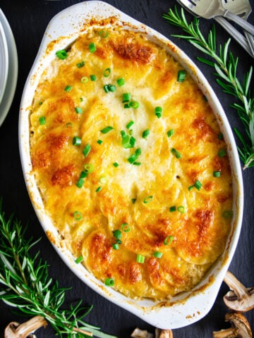 Vertical image of Scalloped Potatoes in a baking dish with rosemary sprigs, forks, and sliced mushrooms nearby