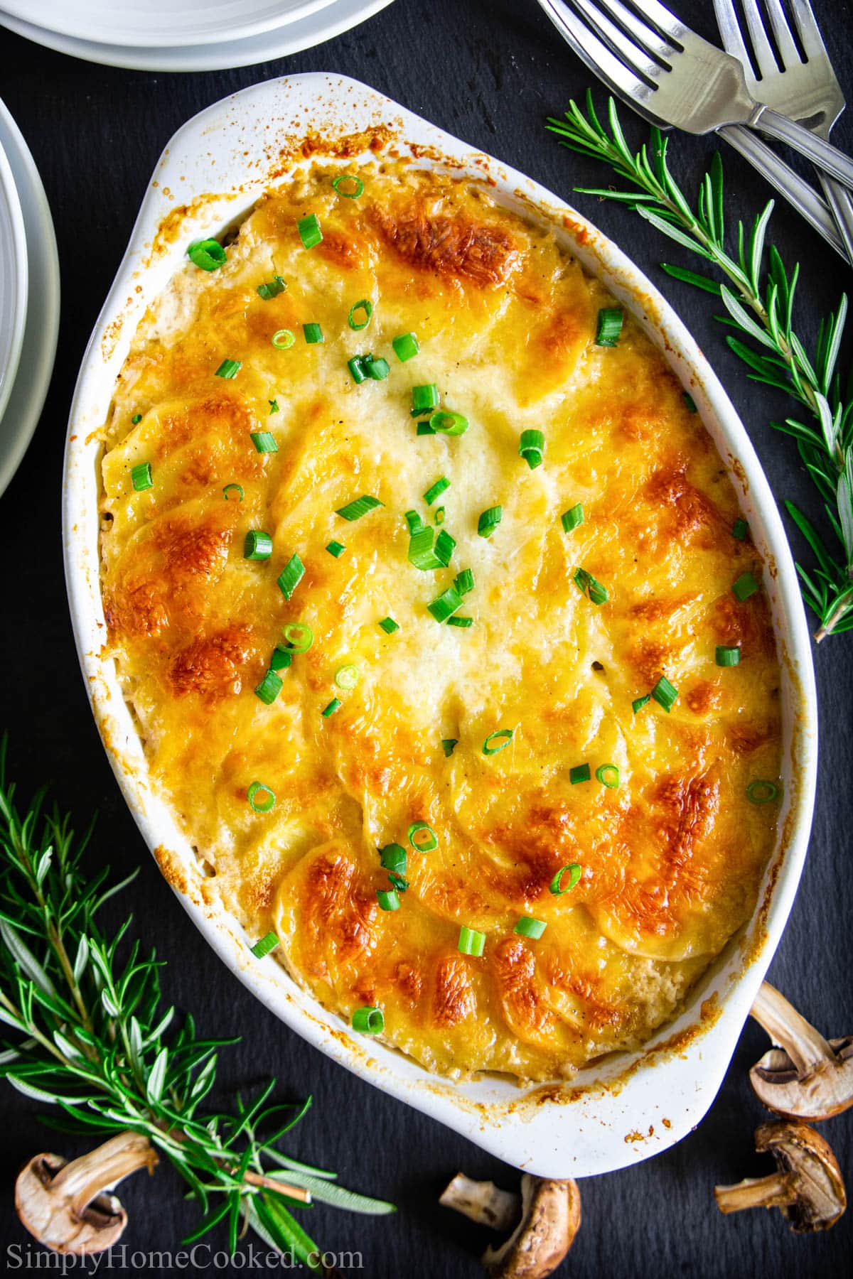 Vertical image of Scalloped Potatoes in a baking dish with rosemary sprigs, forks, and sliced mushrooms nearby