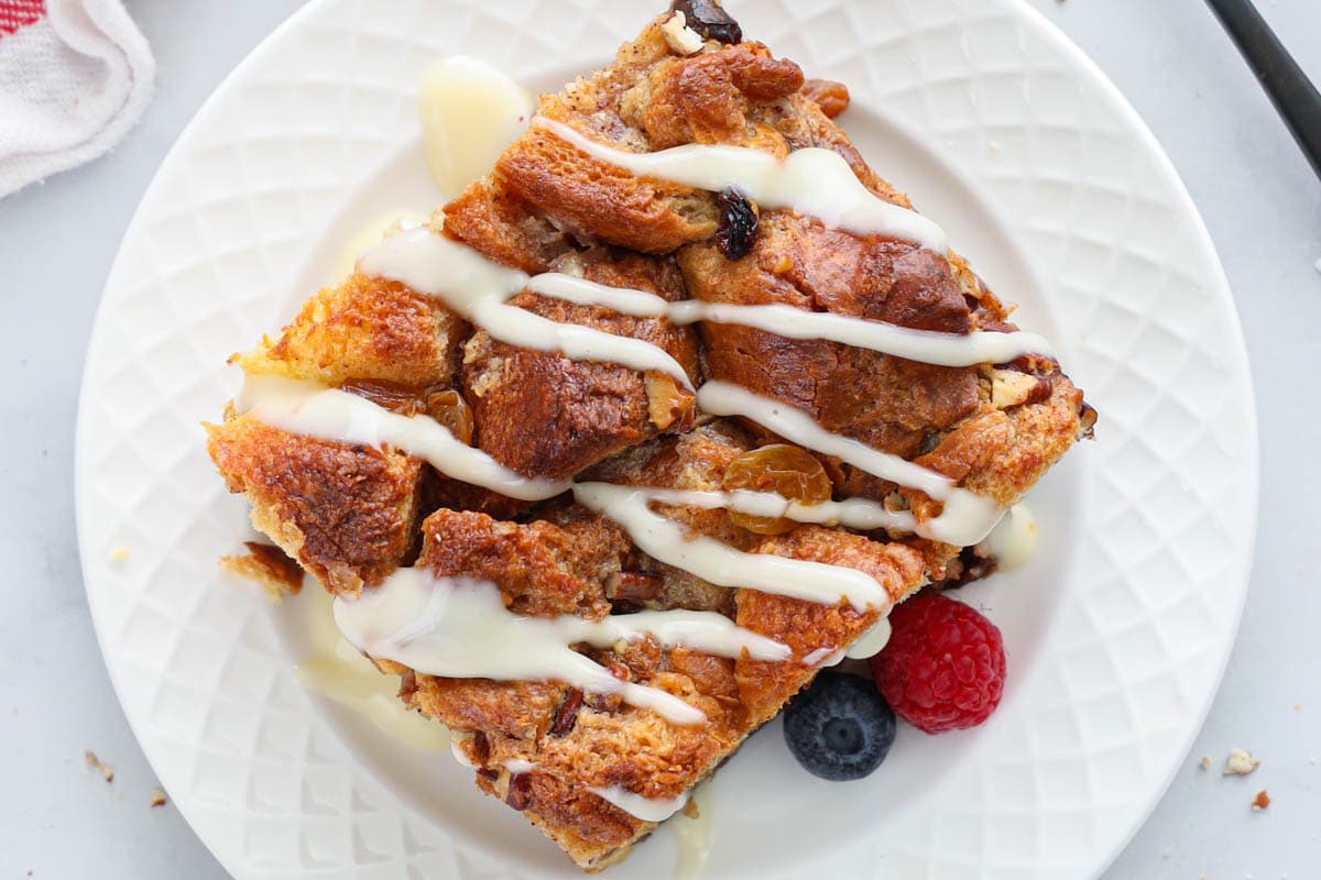 Horizontal overview image of a slice of Bread Pudding drizzled with vanilla rum sauce on a white plate with berries.