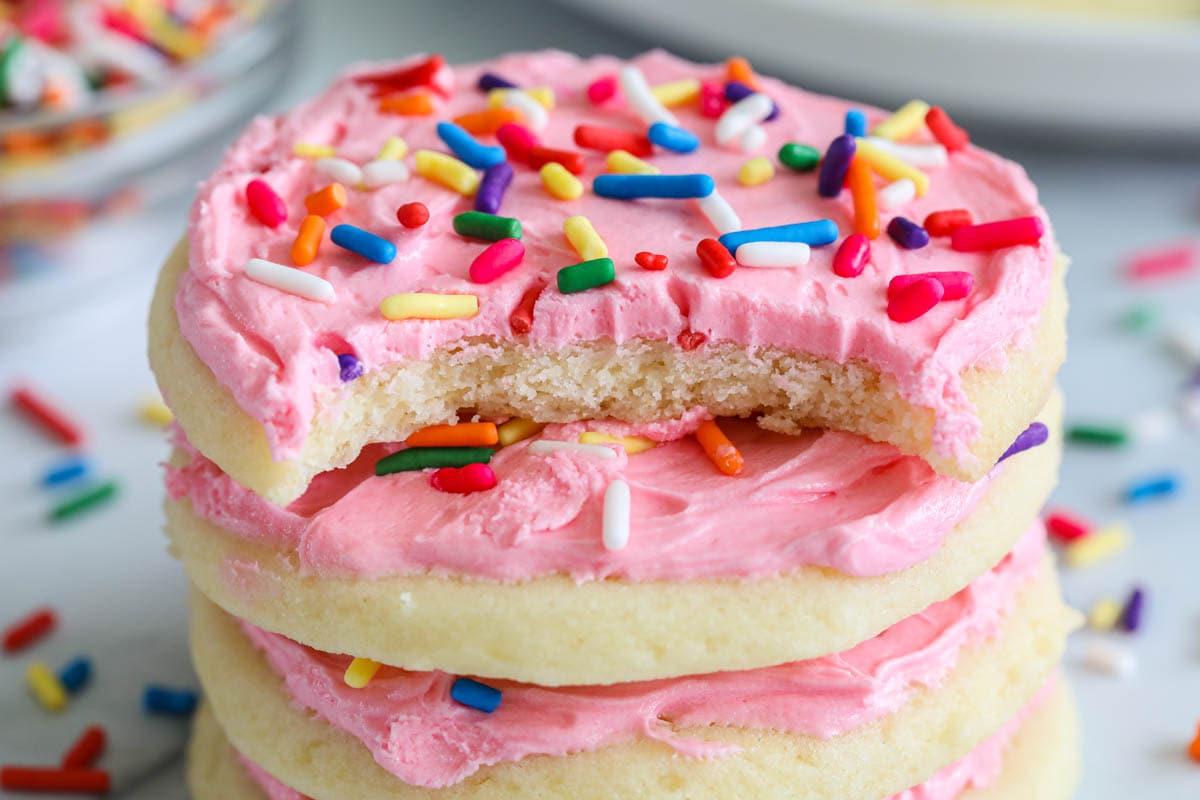 Close up of a stack of Lofthouse Frosted Sugar Cookies with pink frosting and sprinkles, and a bite missing from the top cookie, sprinkles in the background.