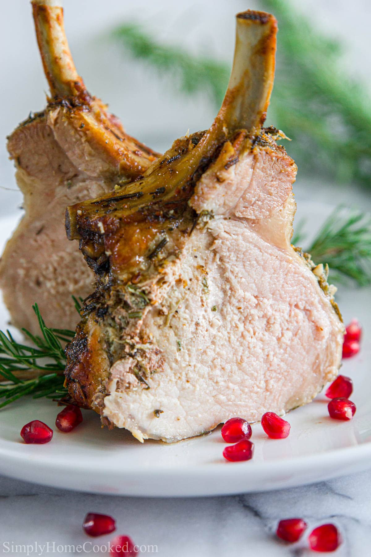 Vertical image of two pieces of Pork Crown Roast on a plate with rosemary and pomegranate seeds