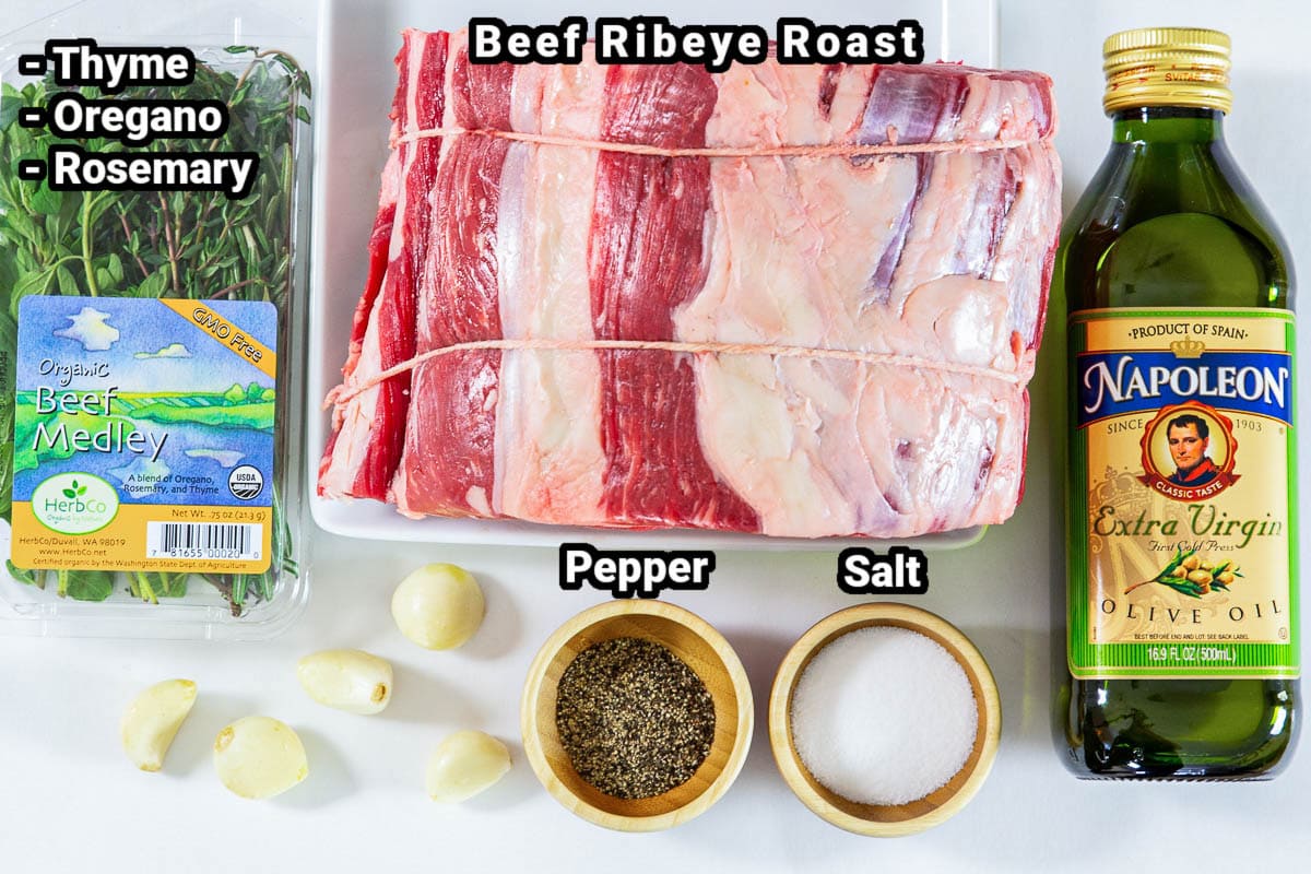 Ingredients for Prime Rib, including beef ribeye roast, olive oil, thyme, oregano, rosemary, salt, pepper, and garlic cloves.