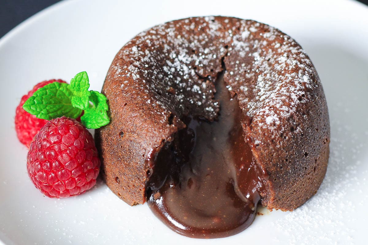 Horizontal image of a split open Chocolate Lava Cake with powdered sugar on top and raspberries next to it