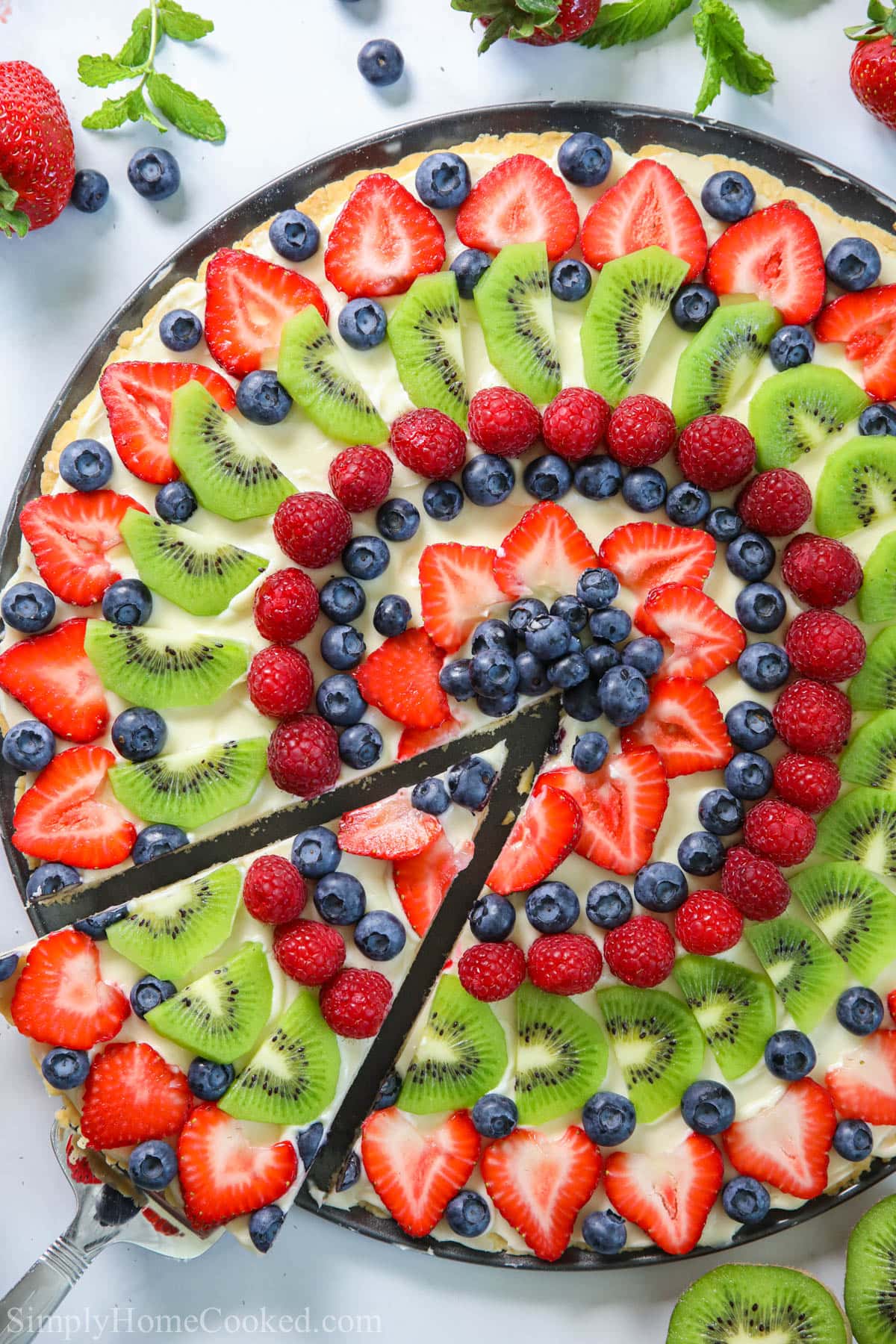 Fruit Pizza with a slice being lifted out and strawberries and kiwis nearby