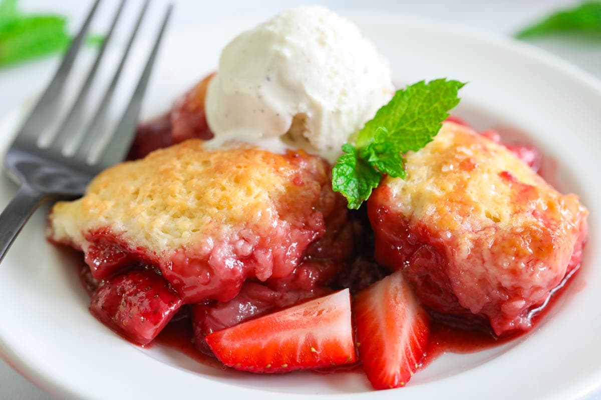 Strawberry Cobbler on a white plate with a fork, ice cream, and fresh strawberries