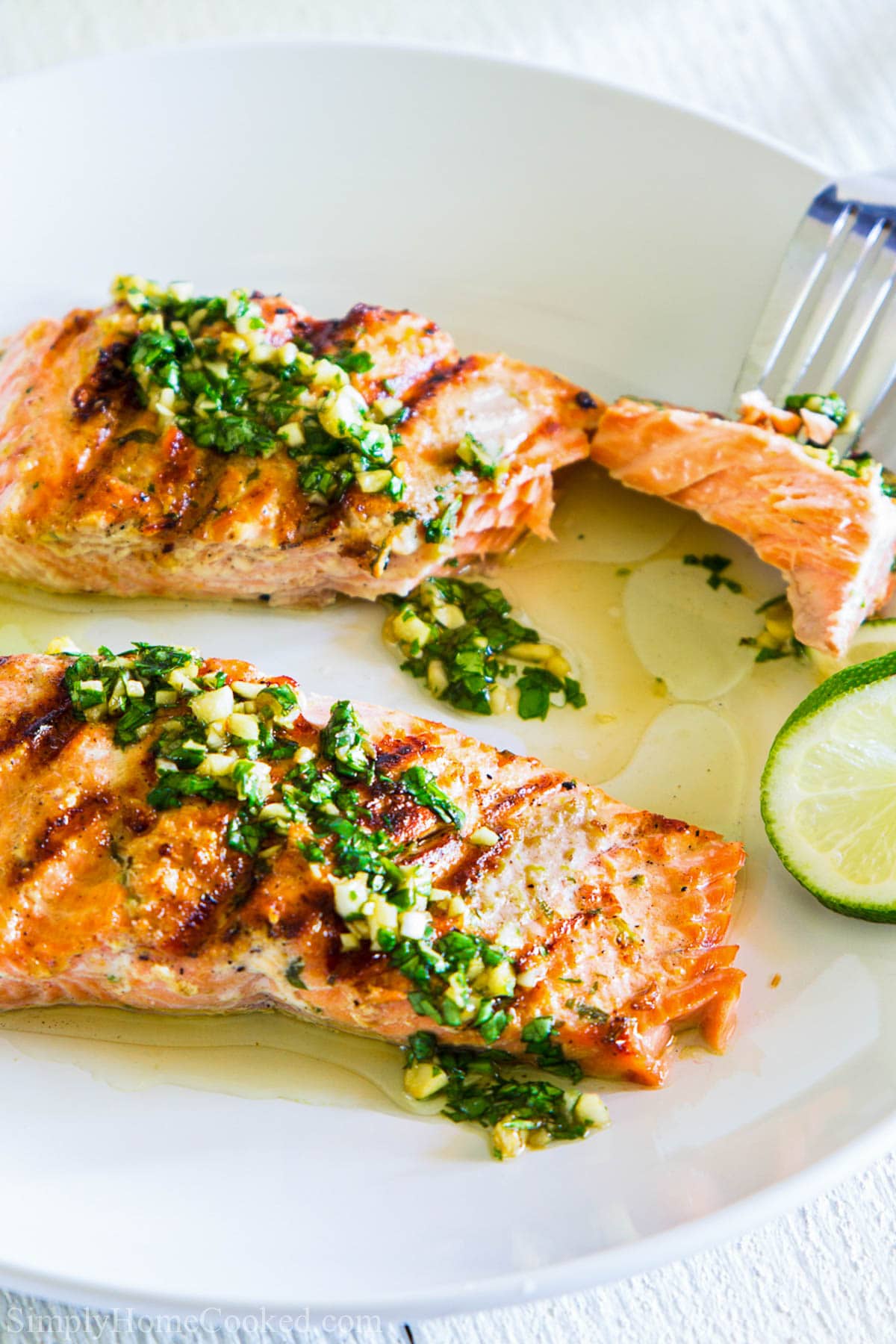 Cilantro Lime Salmon fillets on a white plate with lime, a piece being stabbed by a fork.