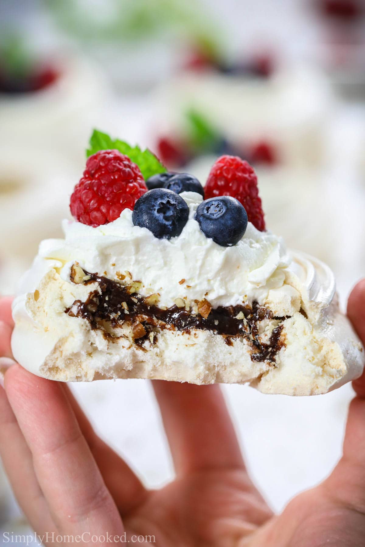 Mini Pavlova with Chocolate and pecans and topped with whipped cream and berries, missing a bite and being held in a hand