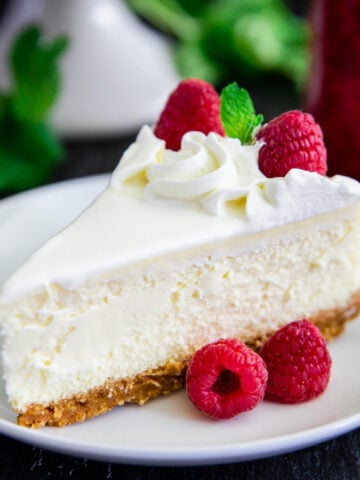 A slice of New York Style Cheesecake topped with whipped cream, fresh raspberries, and mint leaves on a white plate.