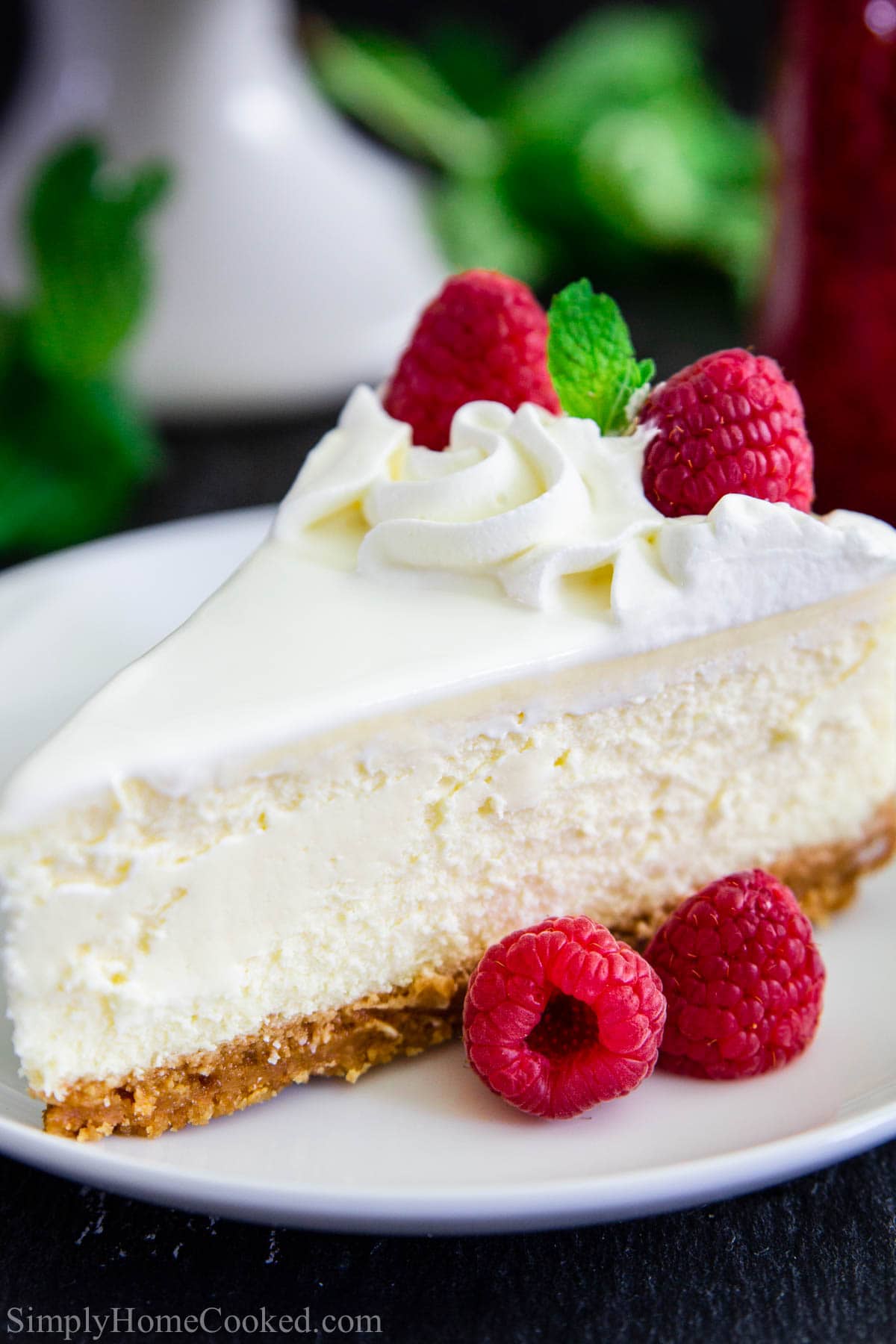 A slice of New York Style Cheesecake topped with whipped cream, fresh raspberries, and mint leaves on a white plate.