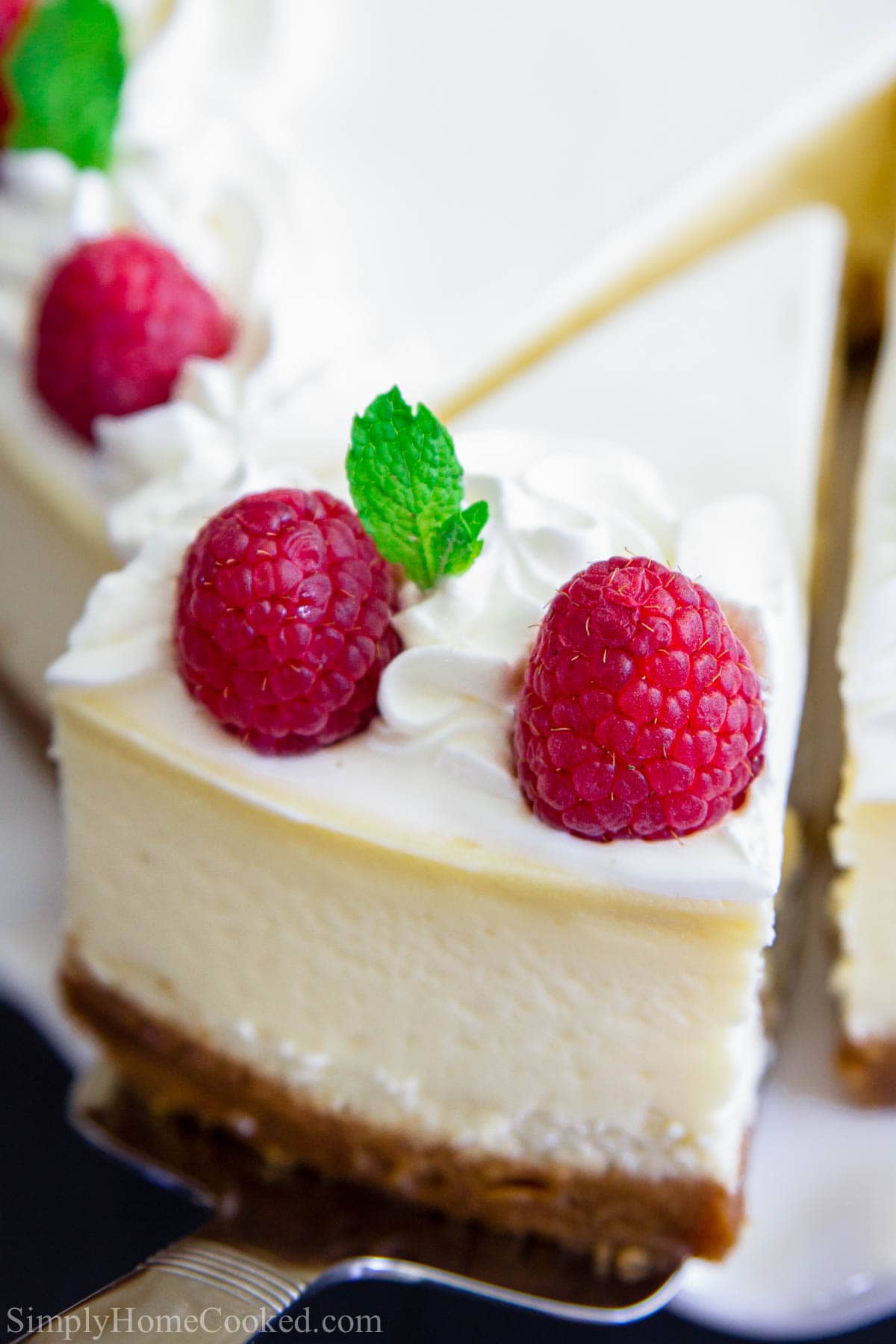 A slice of New York Style Cheesecake being lifted out and topped with whipped cream, fresh raspberries, and mint leaves.