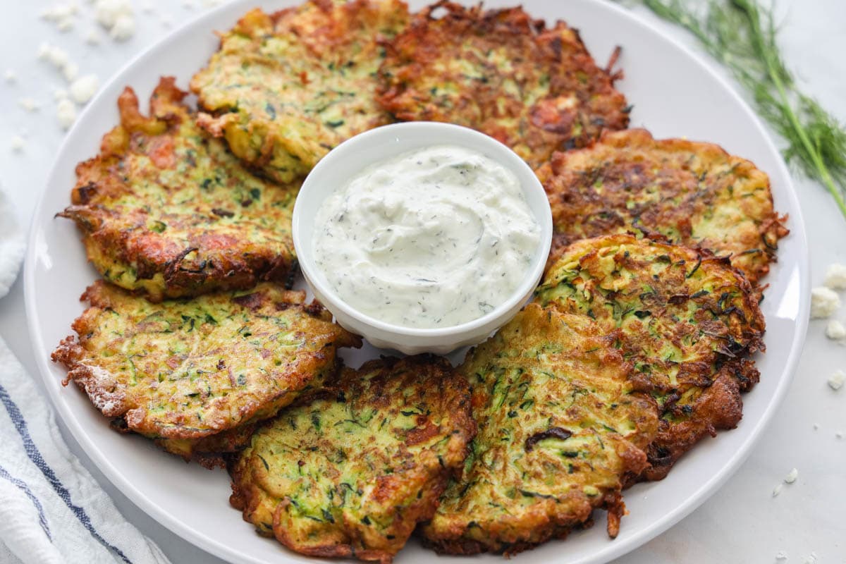 Zucchini are surrounded on a white plate with tzatziki sauce in the middle