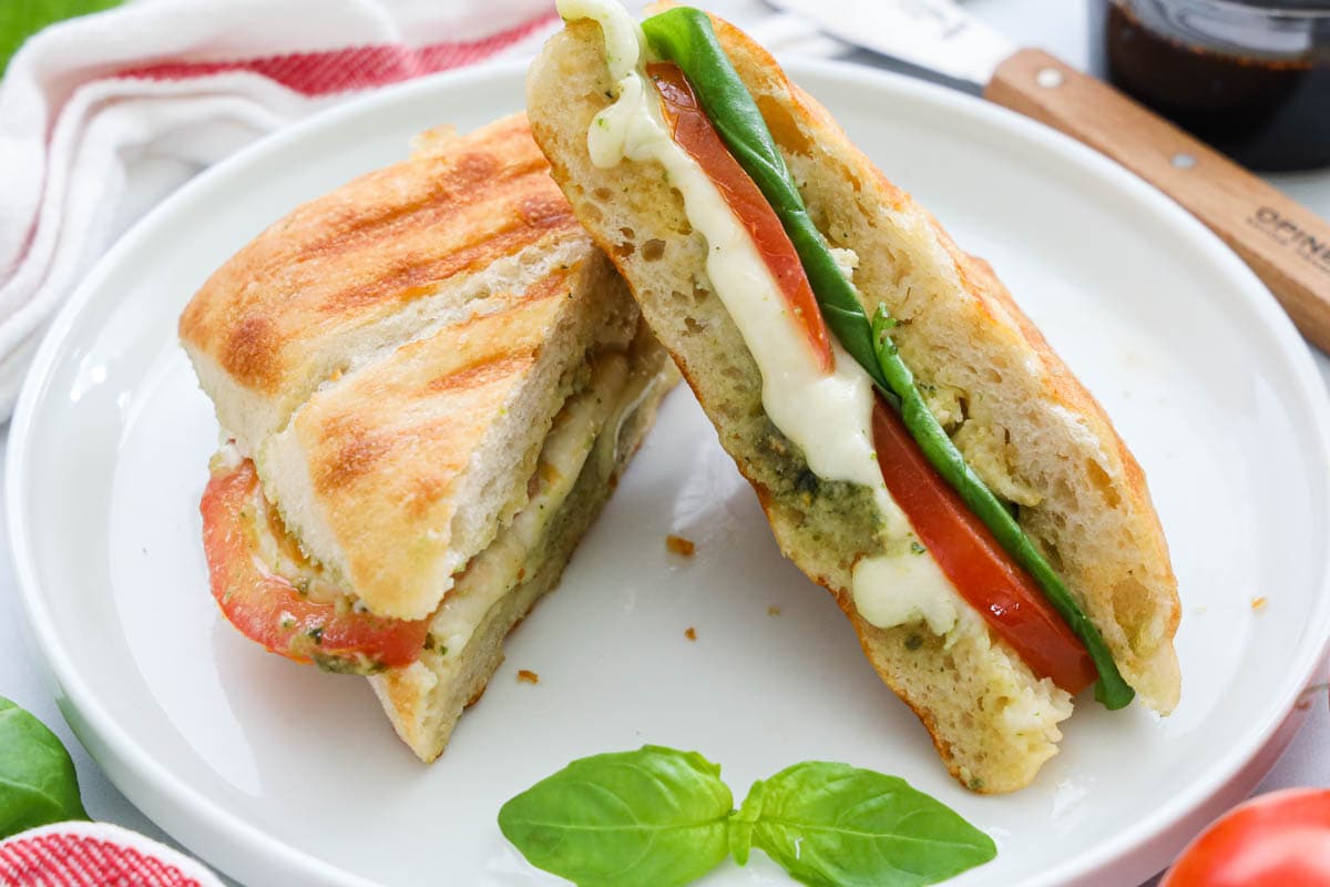 Two halves of a Caprese Sandwich on a white plate with basil leaves.