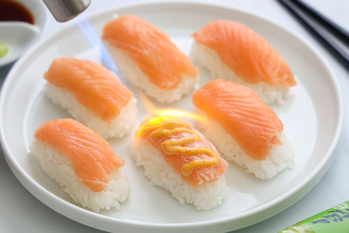 Salmon Nigiri on a white plate, one with spicy mayo on top and being seared by a blowtorch
