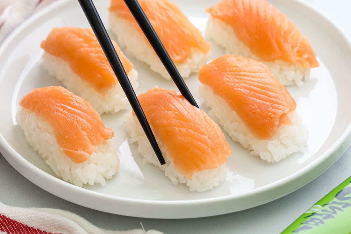 Salmon Nigiri on a white plate, one being lifted by black chopsticks
