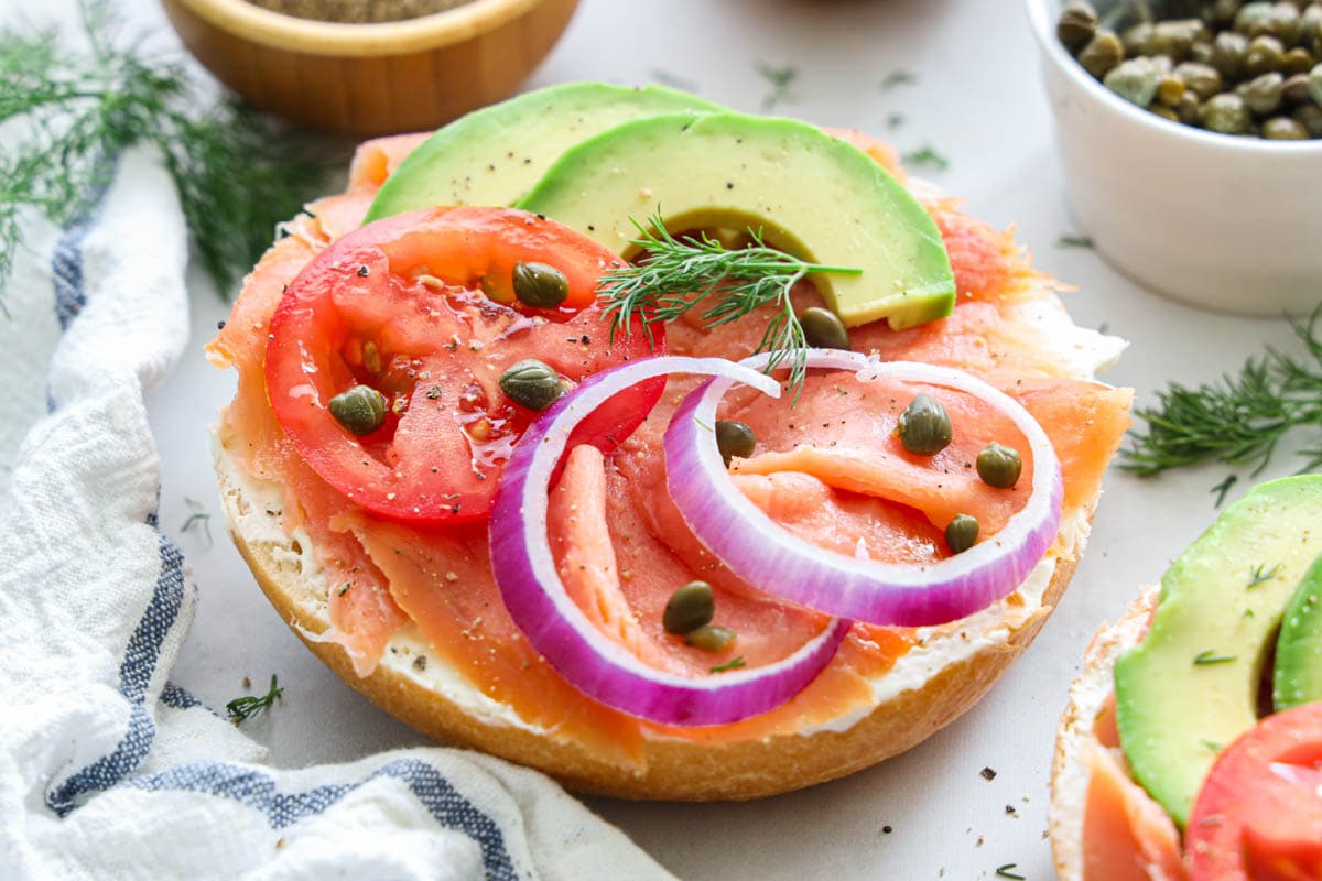 Smoked Salmon Bagel topped with onion, capers, tomato, dill, and avocado, with more nearby.