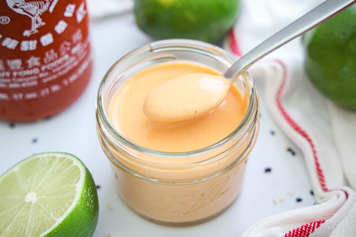 Spicy Mayo in a mason jar with a spoon, and limes and Sriracha nearby.