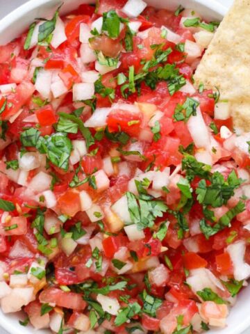 Overhead view of a white bowl of Pico de Gallo with a chip in it and more nearby