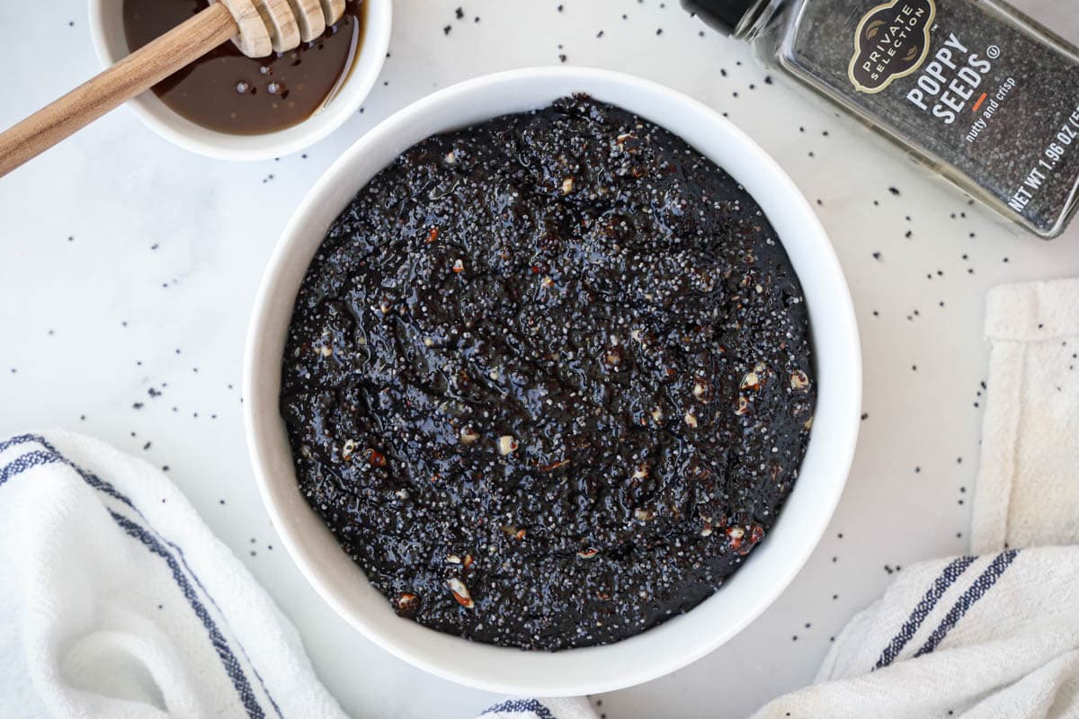 Poppy Seed Filling in a white bowl on a tablecloth with poppy seeds and honey nearby.