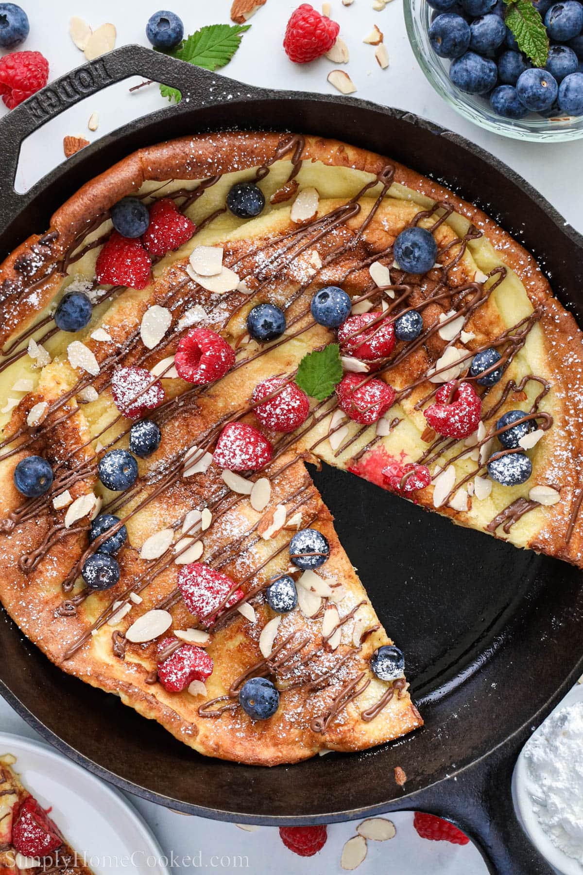 German Pancake in a cast iron skillet topped with Nutella, sliced almonds, berries, and powdered sugar, one slice missing.
