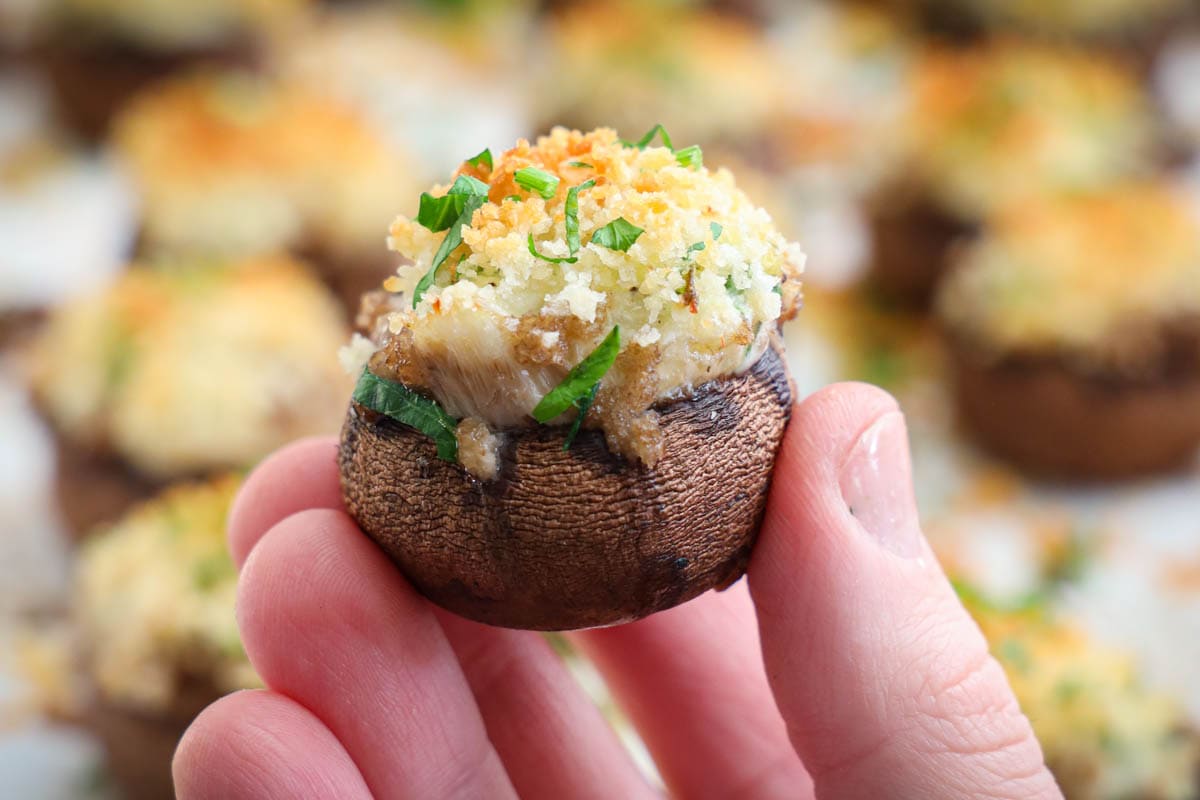 A hand holding a Crab Stuffed Mushroom topped with butter and Panko, browned to perfection and garnished with chopped parsley.