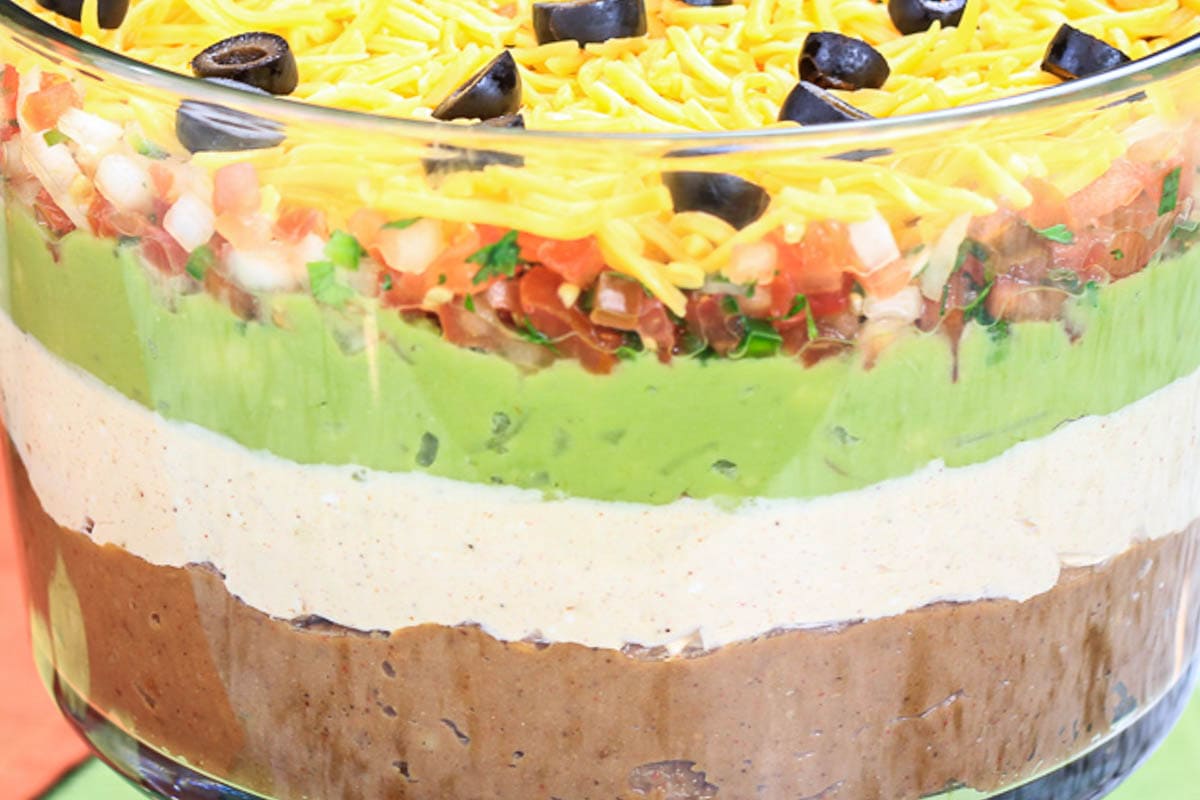 7 Layer Taco Dip in a trifle bowl with beans, sour cream mixture, guacamole, pico de gallo, cheese, and olives.