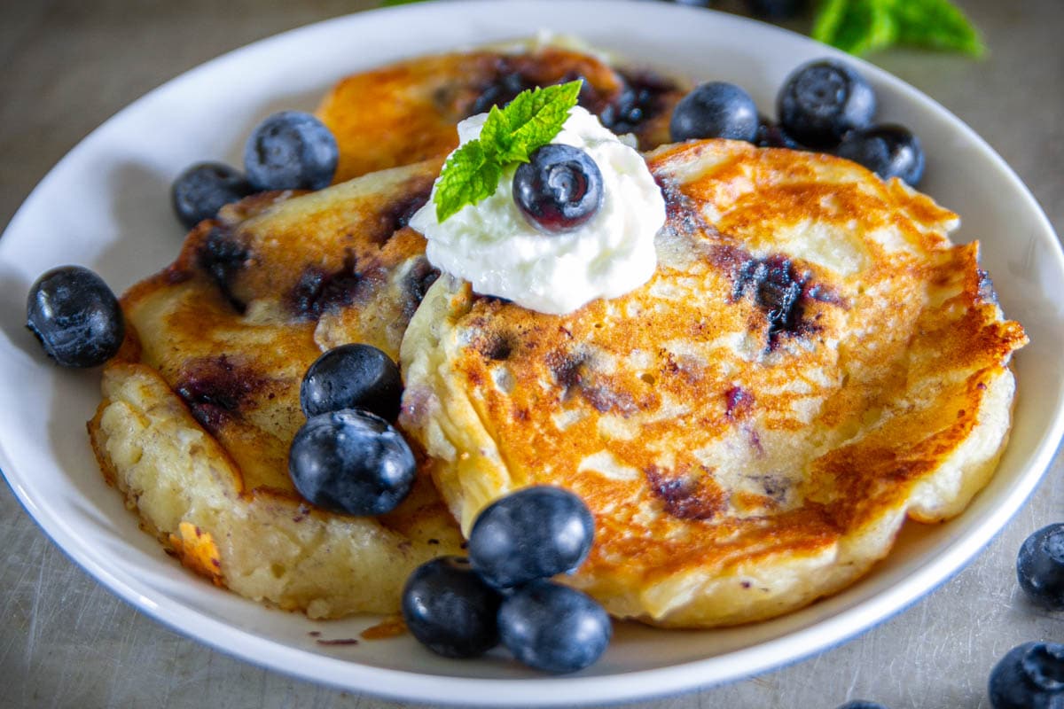 Plate of Blueberry Ricotta Pancakes topped with whipped cream and a mint sprig and more blueberries.