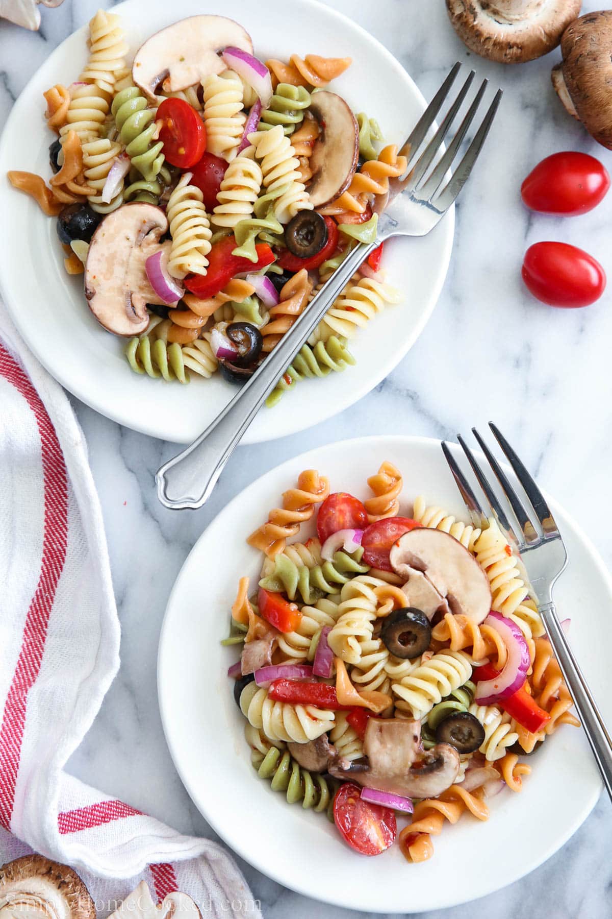 Two plates of Tri Color Pasta Salad with forks and mushrooms and cherry tomatoes nearby.