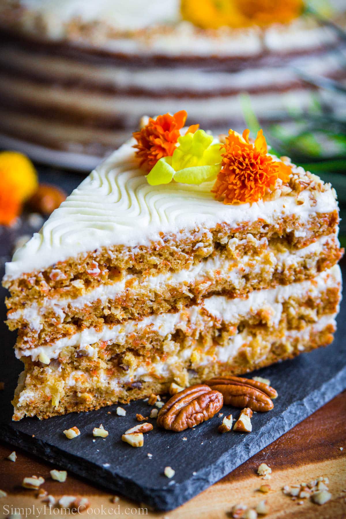A slice of Easy Carrot Cake toped with flowers and pecans next to it. 