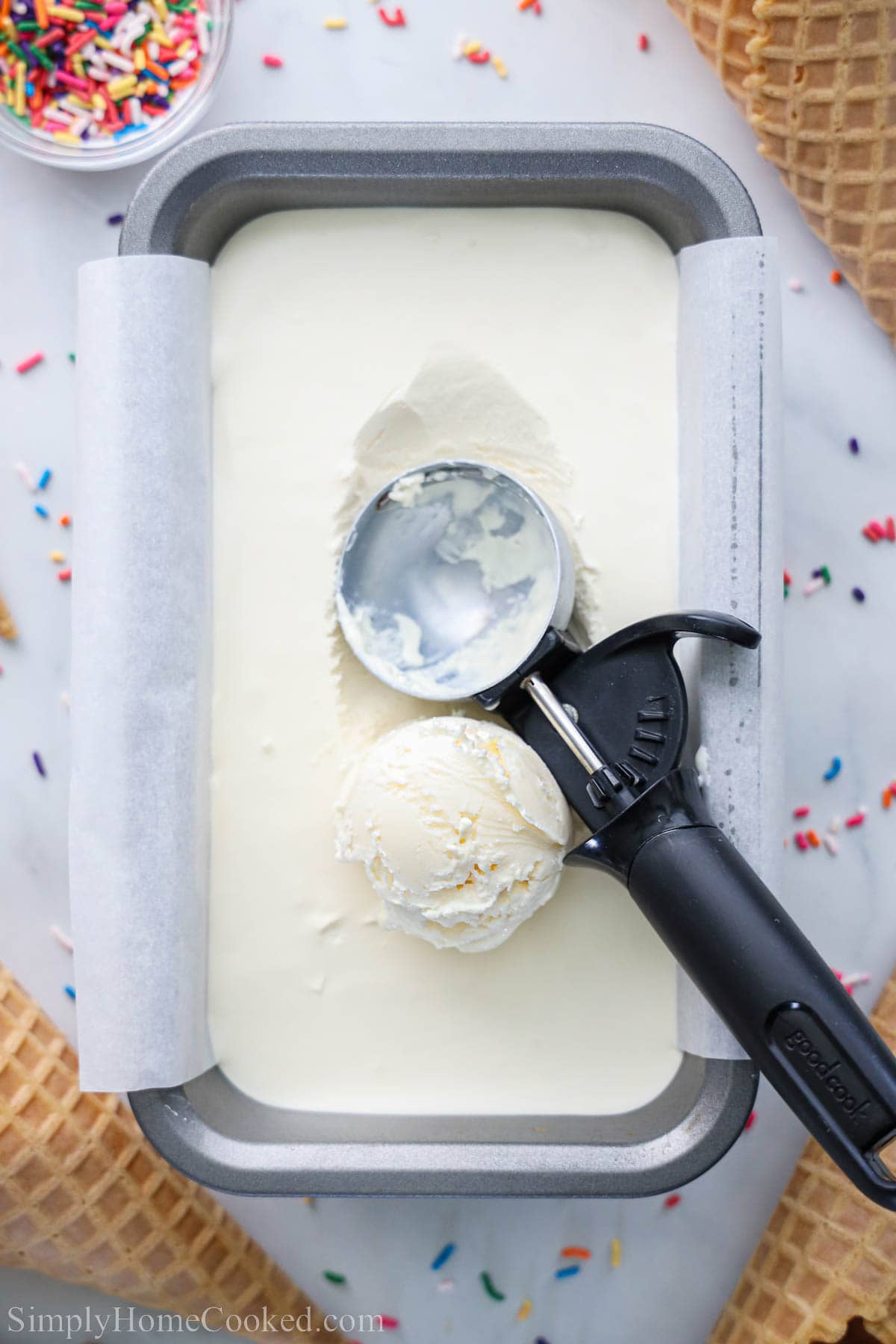 No Churn Ice Cream in a loaf pan with an ice cream scooper and sprinkles and waffle cone nearby.