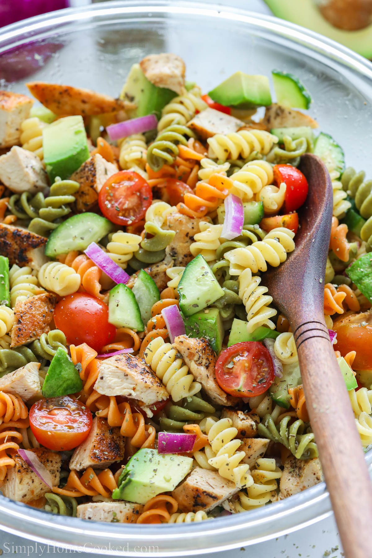 Chicken pasta salad with tomatoes, cucumber, avocado, onion, rotini, and chicken in a bowl with a wooden spoon.