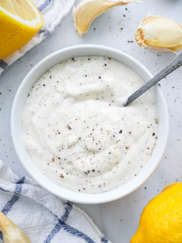 Garlic Aioli in a bowl with a spoon and lemons and garlic cloves nearby.