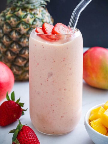 Tropical Smoothie in a glass with a straw and strawberry slices on top, mangos, pineapple, and strawberry nearby.