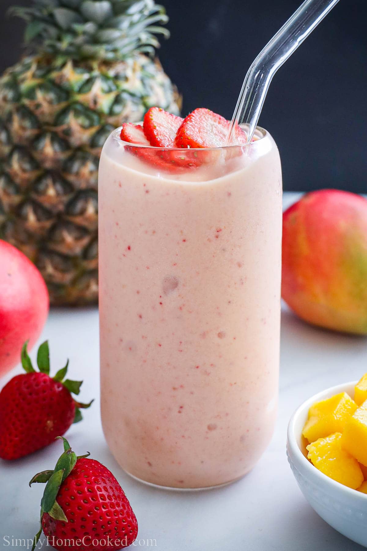 Tropical Smoothie in a glass with a straw and strawberry slices on top, mangos, pineapple, and strawberry nearby.