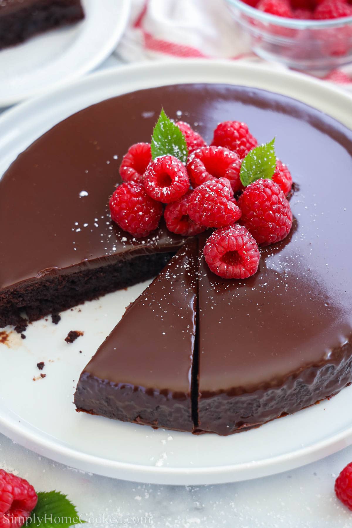 A Flourless Chocolate Cake missing a slice and topped with chocolate ganache, powdered sugar, and fresh raspberries.