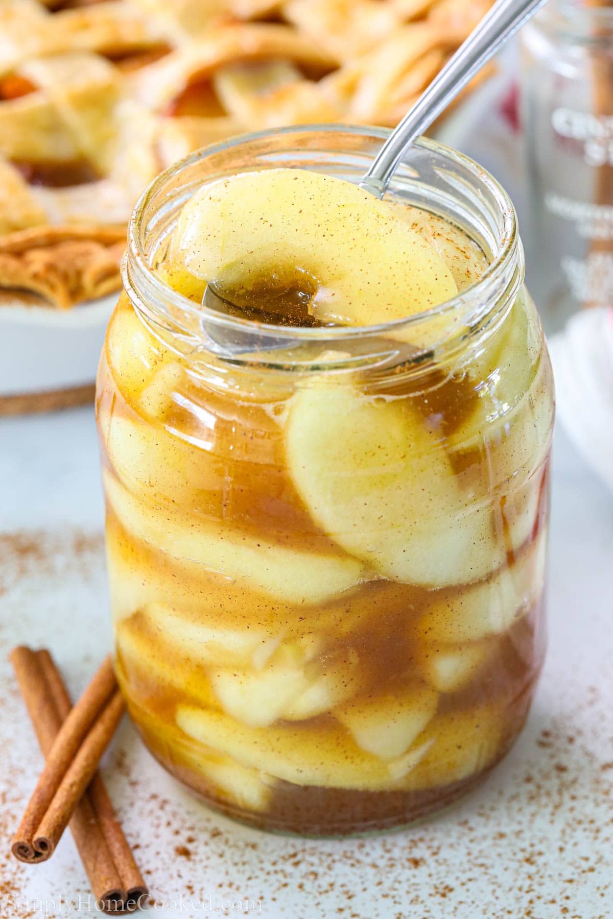 Homemade Apple Pie Filling in a mason jar with a spoon and cinnamon sticks nearby.
