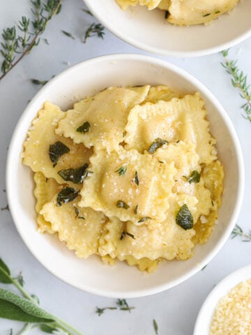 Butternut Squash Ravioli in a white bowl with fresh sage and thyme nearby.