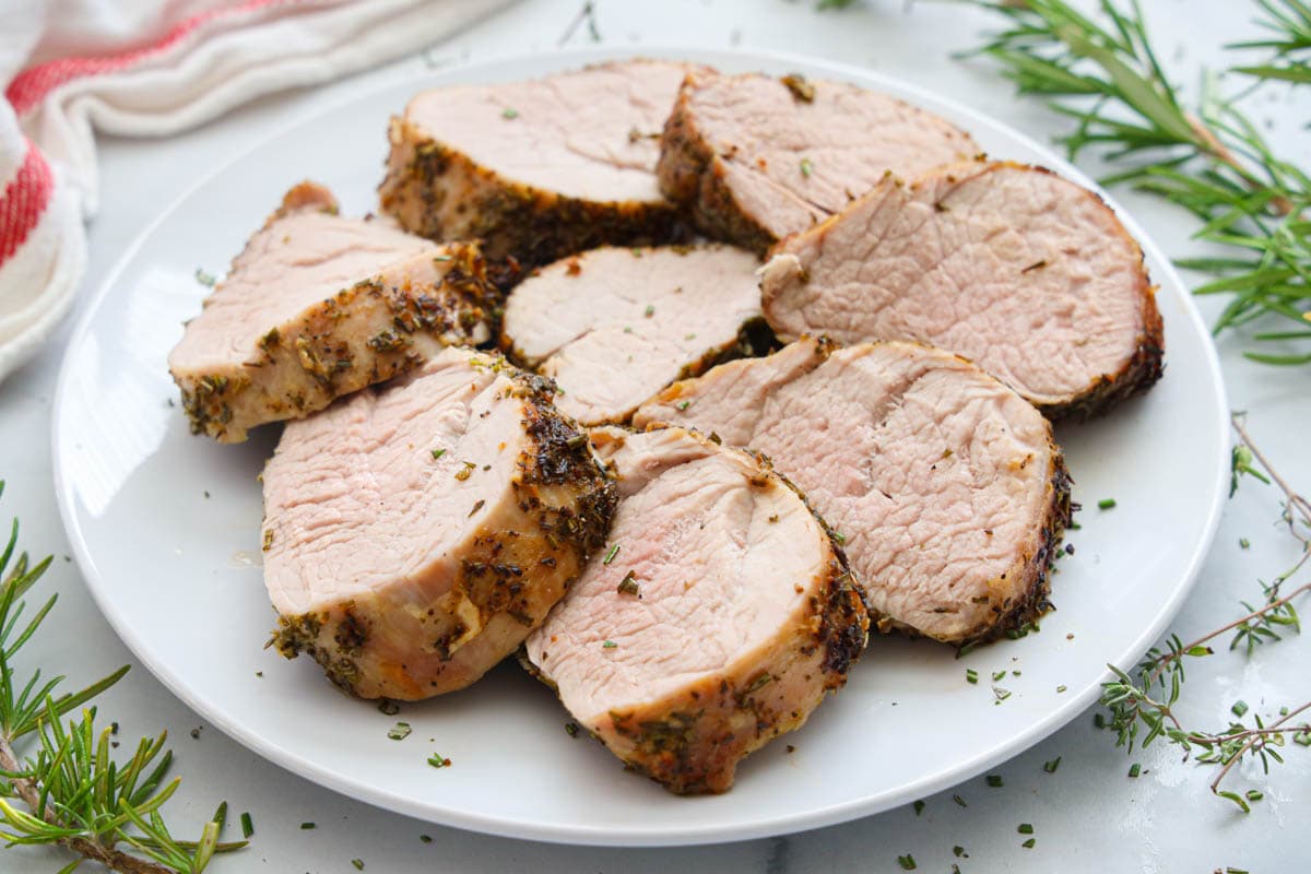 Air Fryer Pork Tenderloin sliced on a white plate with fresh rosemary and thyme nearby. 