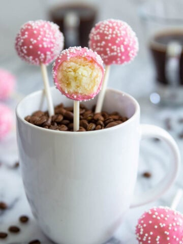 Vanilla Cake Pops covered in pink chocolate coating and sprinkles, then placed in a mug with coffee beans.