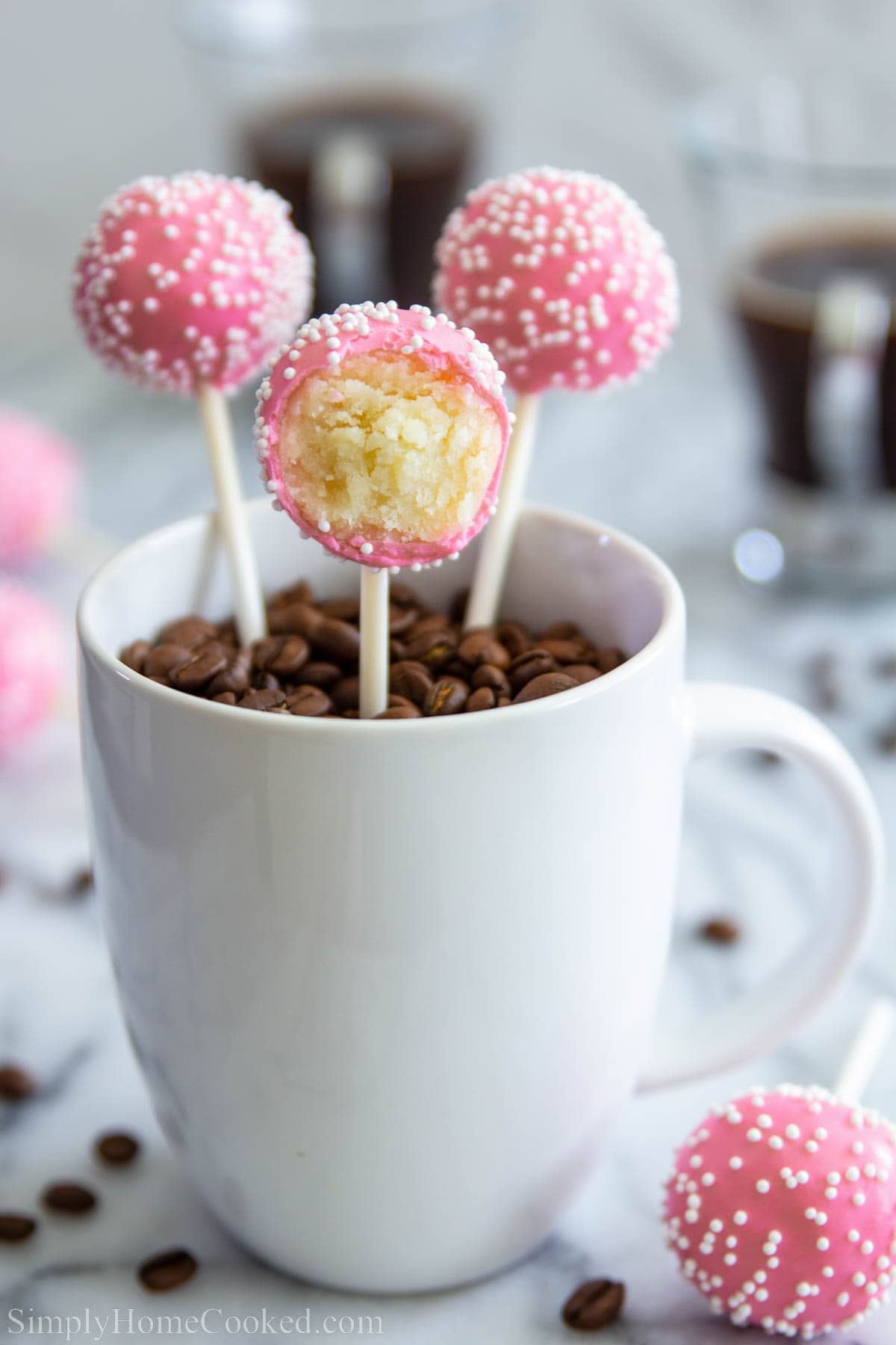 Vanilla Cake Pops covered in pink chocolate coating and sprinkles, then placed in a mug with coffee beans. 