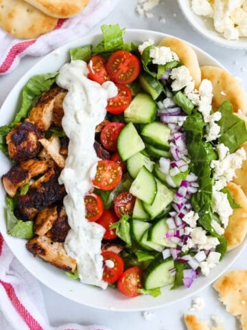 Overview of Chicken Shawarma Bowls, topped with tzatziki sauce, tomatoes, cucumbers, onion, feta cheese, and pita bread.