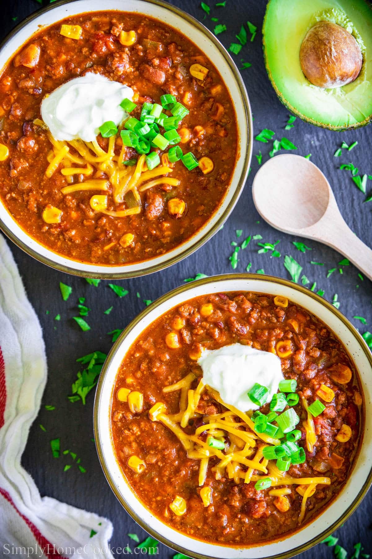 Easy Beef Chili in a bowl with cheese, green onions, and sour cream, and avocado on the side.